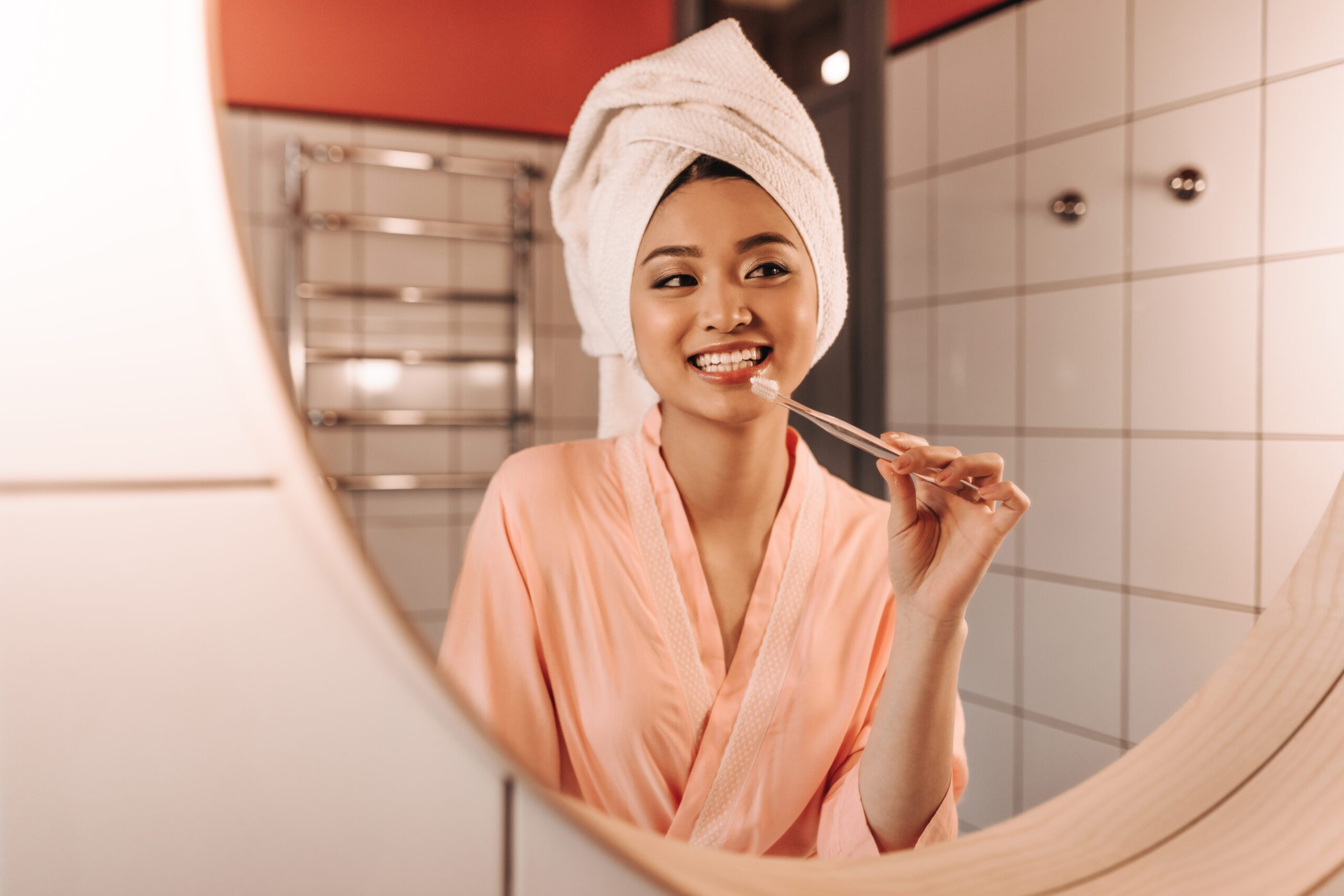 Brown-eyed girl in white towel is brushing her teeth. Woman looking in bathroom mirror